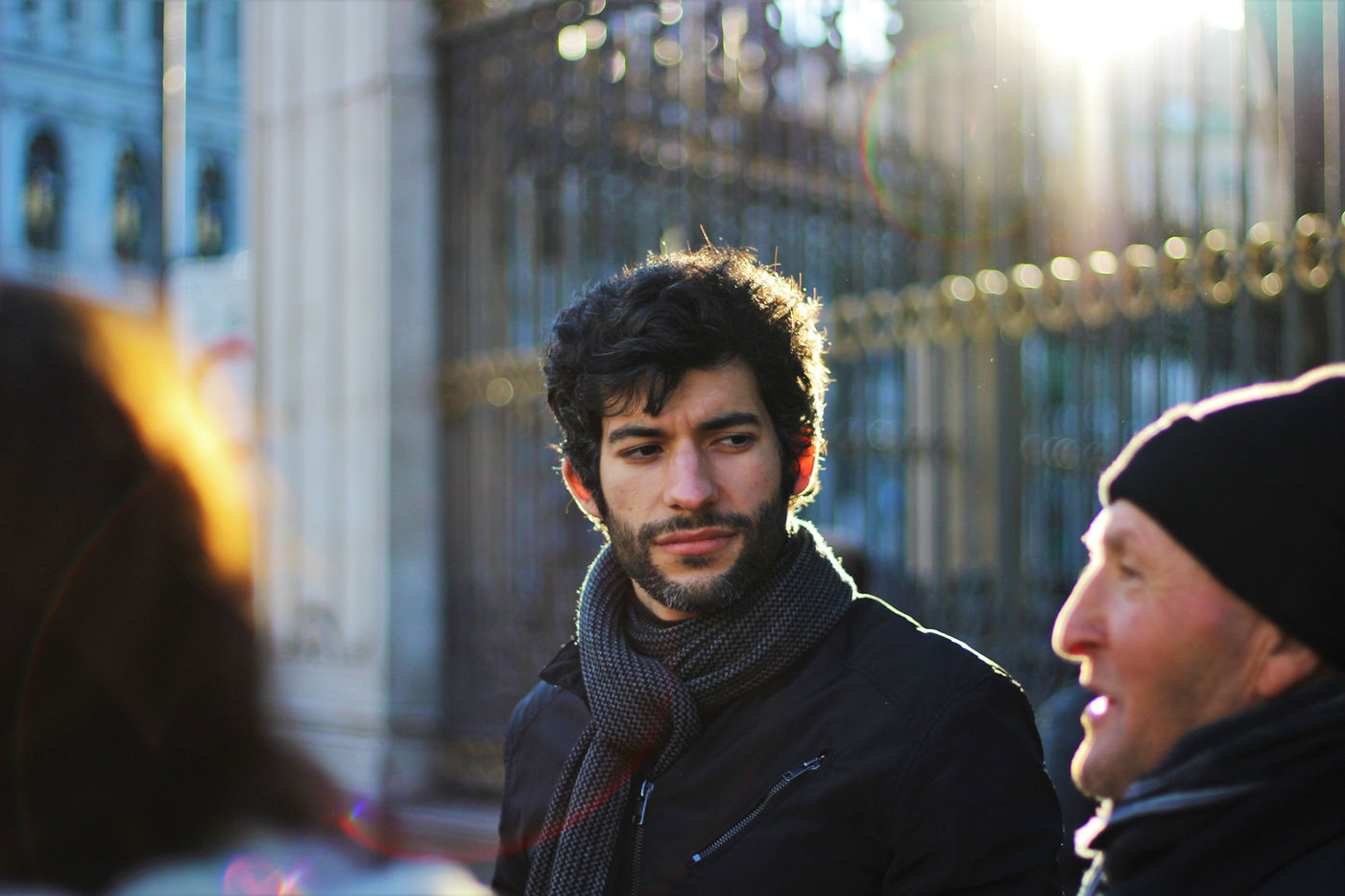 Portrait of a man speaking with two others in front of a metal gate in a metropol.