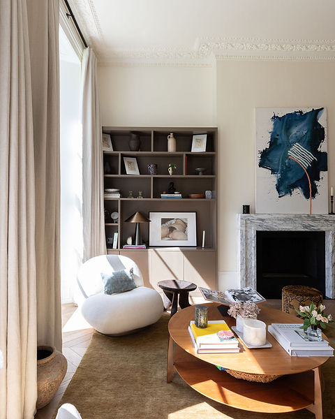 Lounge area with tall open windows. Bespoke joinery in a warm brown sits in the corner decorated with pictures, vases and books complimenting the warm wood coffee table and marble dire place