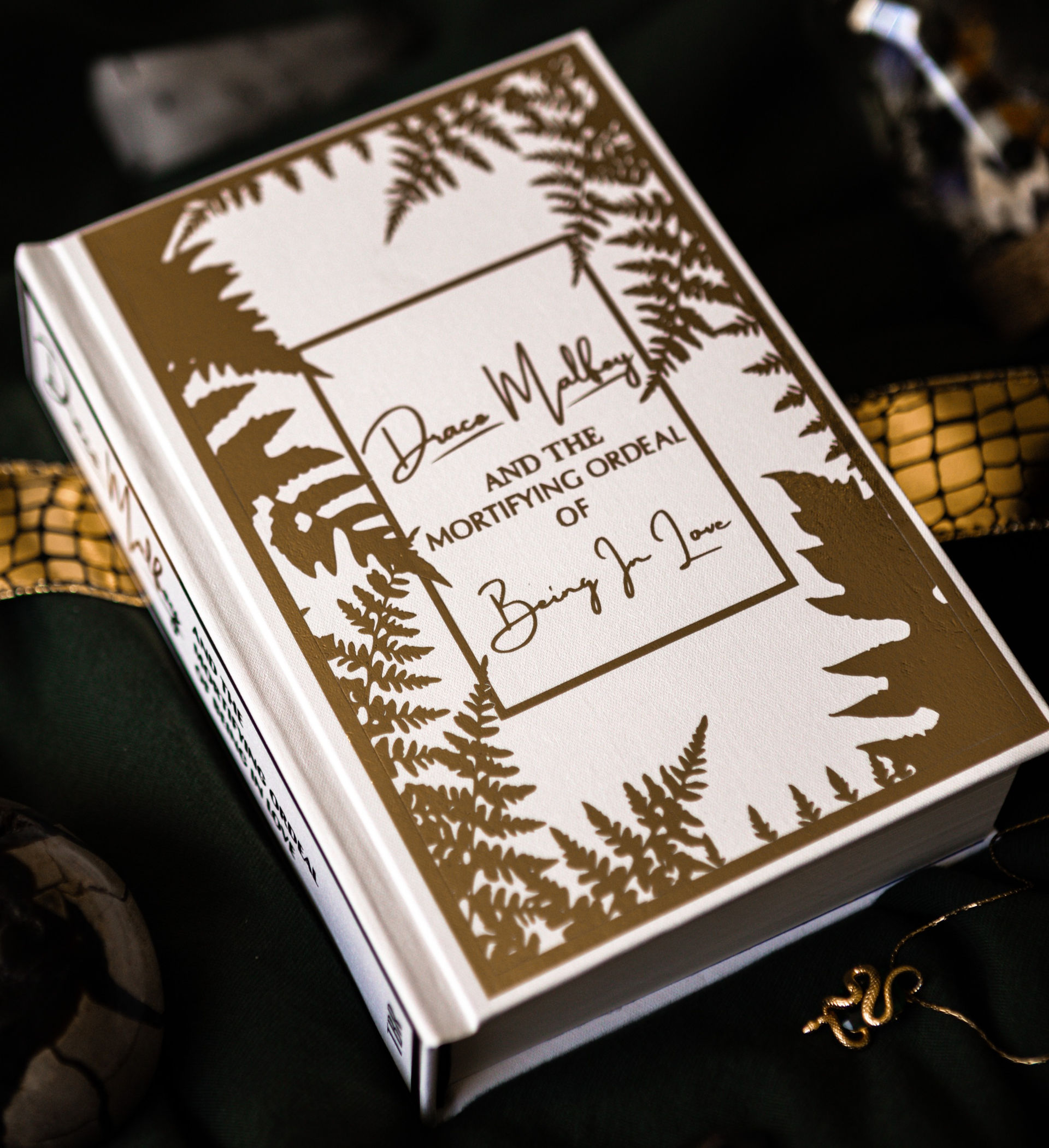 A white hard cover book with gold details including the title of the book and fern leave silhouettes inside of rectangular boarders. 