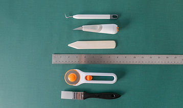 Book binding tools on a green backdrop. From top to bottom: grey and white weeding tool, grey, white and orange blade, white bone folder, silver ruler, white and orange rotary blade, and a grey and black solicone paintbrush.
