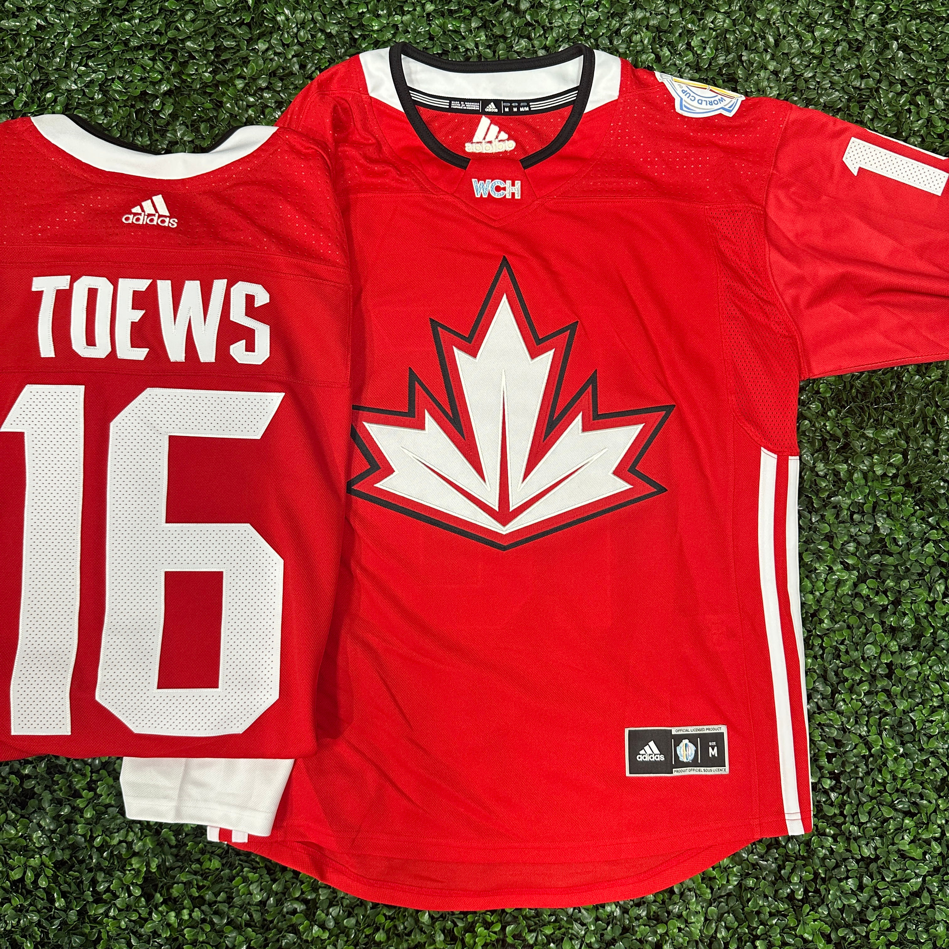 A Red  WCH Hockey Jersey that has a White Maple Leaf at the front and white srtripes at the ribs of the jersey. The same jersey is folded next to it displaying the backside of it that has "TOEWS" AND "16" directly under.
