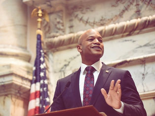 A photograph of Governor Wes Moore dressed in a dark suit, patterned tie, and white shirt stands at a podium in an ornate government chamber with marble walls and an American flag in the background, gesturing with one hand while speaking.