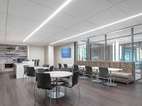 A modern office break room with a mix of round and square tables, wood chairs and a colorful booth style bench.