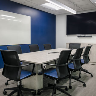Small conference room with navy accent wall, large whiteboard, wall-mounted video screen, white oval table, and six blue task chairs.