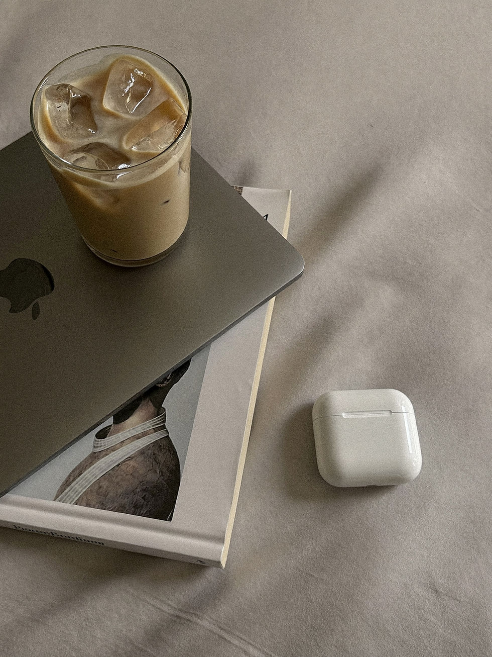 Laptop on top of a magazine next to airpods and an iced coffee.