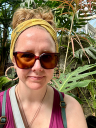 Caitlin with a mustard yellow headband and sunglasses in front of large plants