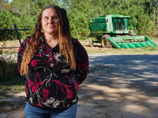 Mujer seria de pie junto a una cosechadora verde en un camino rural, con bosque de fondo y blusa negra de flores.