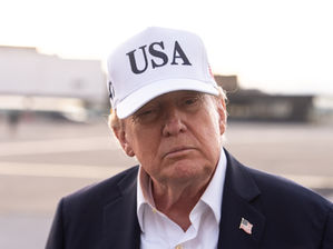 Primer plano de Donald Trump con gorra blanca USA y traje oscuro, gesto serio, al aire libre con fondo desenfocado.