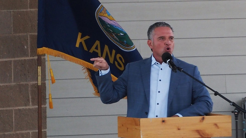 Hombre de traje habla en un podio y señala, con micrófono y bandera de Kansas al fondo.