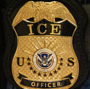 Placa dorada de agente de ICE con sello del Departamento de Seguridad Nacional sobre fondo oscuro.