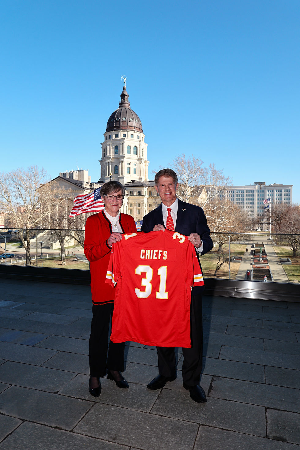 La gobernadora Laura Kelly posa junto al presidente de los KC Chiefs sosteniendo la camiseta del equipo.