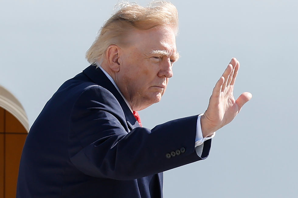 Trump saluda con la mano desde un avión, con traje oscuro y corbata roja, bajo un cielo claro.