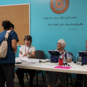 Mujer se registra en una mesa de votación con tres voluntarias; pared turquesa con Proverbios 4:11 y mostradores electrónicos.