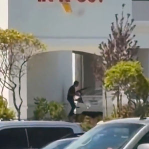 Entrada de In-N-Out con letrero rojo; una persona empuja un carrito frente al local, con autos y arbustos al frente.