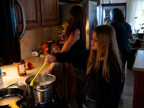 Niña remueve una olla en la cocina; dos adultos cocinan detrás, con luz cálida y ambiente familiar tranquilo.
