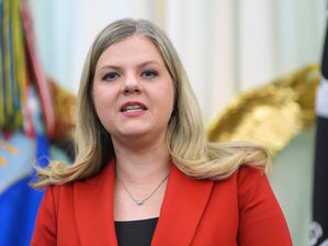 Mujer rubia con blazer rojo habla ante banderas desenfocadas; expresión seria en un acto oficial.