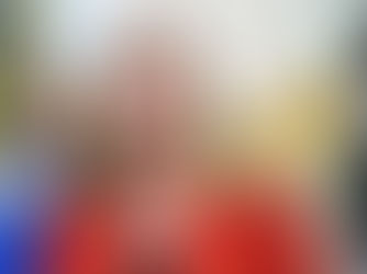 Mujer rubia con blazer rojo habla ante banderas desenfocadas; expresión seria en un acto oficial.