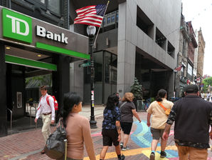 Peatones caminan por una calle urbana junto a un TD Bank; banderas de EE. UU. ondean y el ambiente es animado.