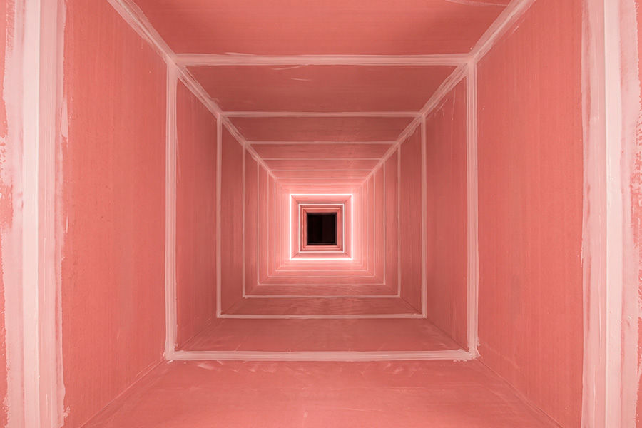 Uma imagem de perspectiva em túnel de incêndio profundo e simétrico, com paredes de drywall resistente a fogo. As juntas entre os painéis de parede e o teto e o chão são visivelmente acabadas com fita adesiva e massa corrida de cor clara, criando um padrão de linhas verticais e horizontais em toda a extensão do túnel. O túnel se estende por um longo caminho, com a estrutura quadrada de drywall se tornando progressivamente menor e mais definida ao longe, criando uma sensação de profundidade e repetição rítmica. No centro da imagem, na extremidade do túnel, há uma pequena luz brilhante e focada, emoldurada por uma pequena e escura abertura quadrada, que serve como o ponto de fuga da perspectiva. A iluminação é uniforme e suave nas paredes, enquanto o centro é iluminado pela luz focada.