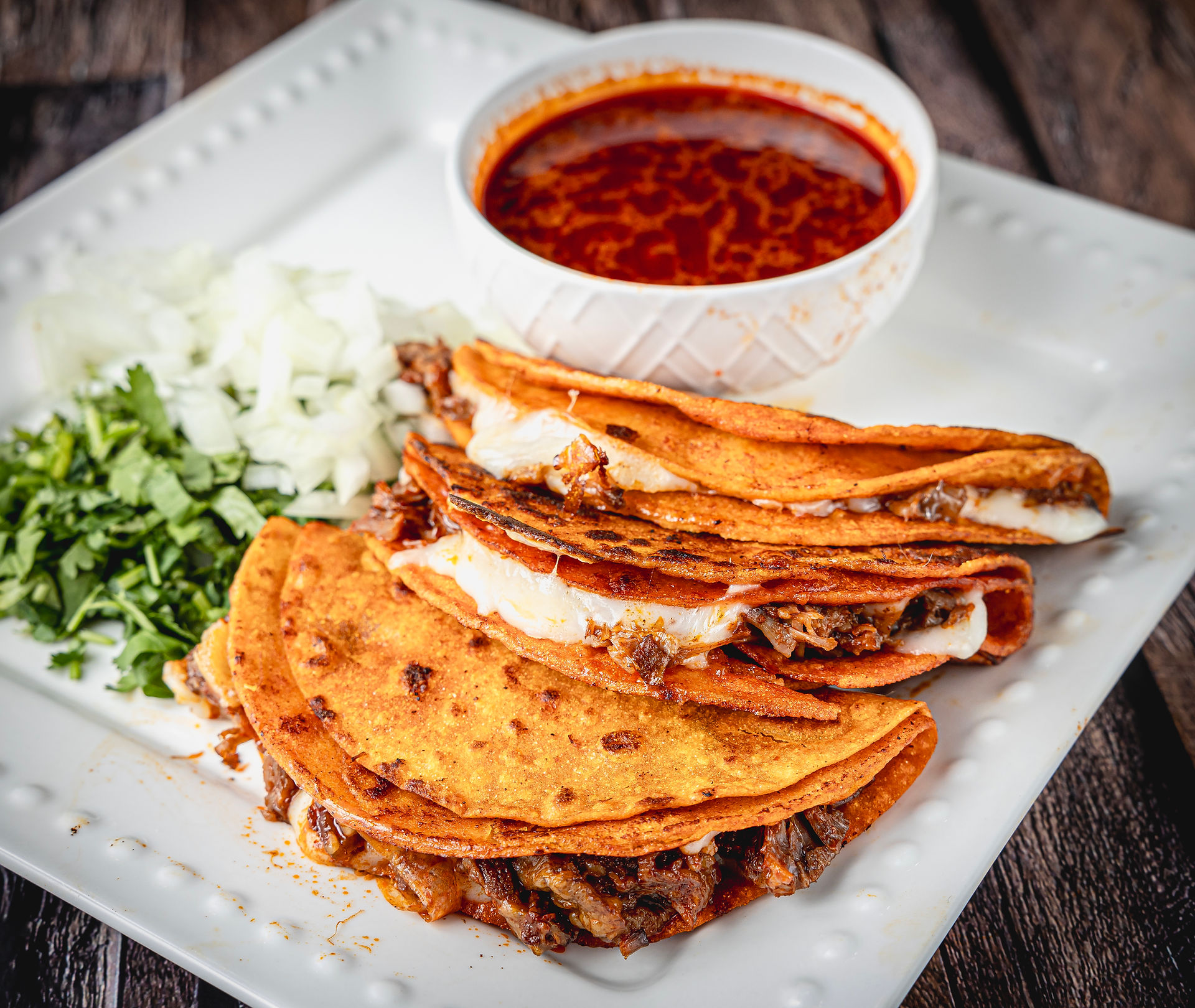 Three grilled quesabirria tacos with onions, cilantro, and consomé on the side