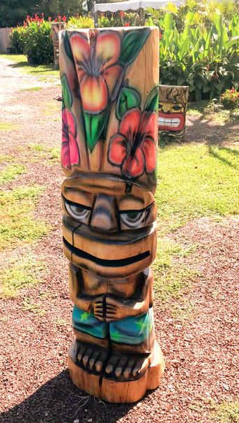 Tiki Murph carved pine full body round tiki in painted shorts and multi colored flowers on top.