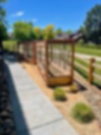 Sunny suburban yard with wooden fenced garden beds beside a sidewalk, green lawn and trees under a clear blue sky.