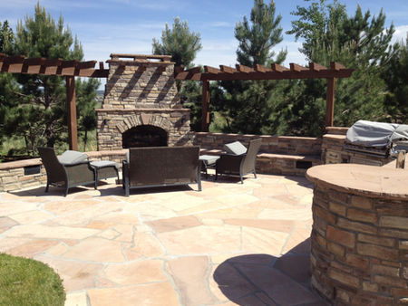 Outdoor patio with stone fireplace, pergola, and wicker furniture. Grill and table nearby. Surrounded by pine trees under a blue sky.