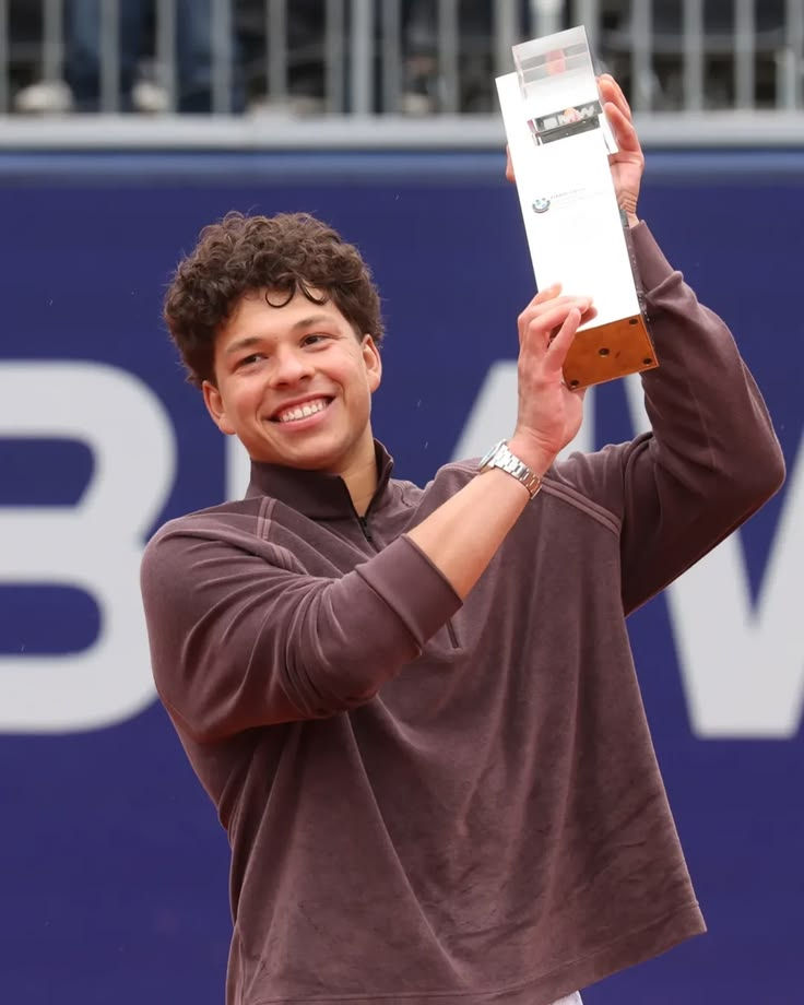 Smiling person Ben Shelton holds up a rectangular trophy in a sports setting, wearing a brown jacket. Background features a blue banner with text.