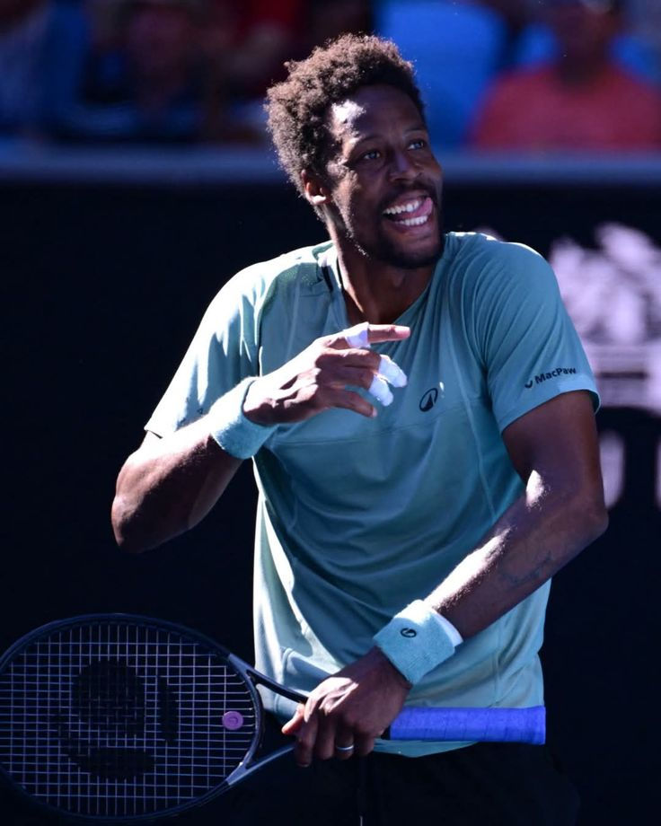 Gael Monfils in a green shirt smiles while holding a racket on a court. Blue wristbands and vibrant backdrop enhance the lively scene.