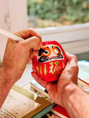 Main d'homme peignant l'oiel d'un daruma rouge, symbole de chance et de bonne fortune