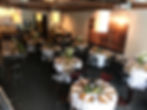 Elegant dining setup with round tables and white cloths, adorned with flowers and candles. Wall displays sunset vineyard art, creating a warm atmosphere.