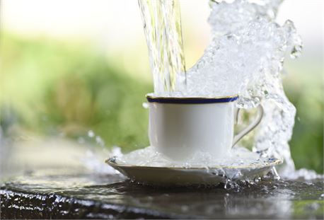 Water overflows a white cup and saucer set outdoors, set against a blurred green background, creating a dynamic and refreshing scene.