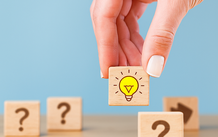 A hand places a wooden block with a glowing light bulb icon among blocks with question marks, symbolizing finding solutions o