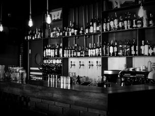 Dimly lit bar in Bangalore with shelves of liquor bottles, hanging lights, beer taps, glassware, and coffee machines. Black and white ambiance.