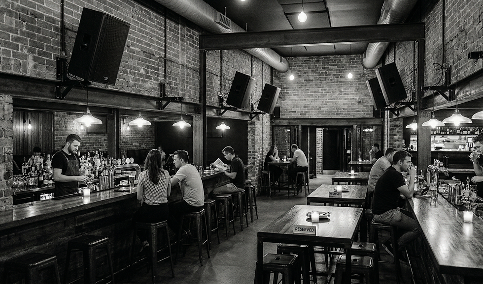 A grayscale, hyper-realistic wide shot of a modern, industrial-style restro-bar. The interior features exposed brick walls, dark wooden bar counters, and visible ceiling ventilation ducts.
To the left and right, bartenders are seen preparing drinks behind the bar, while a few patrons sit on stools or at wooden tables throughout the space. Several large, professional-grade black speakers are mounted to the brick walls and structural beams, angled toward the seating areas. The atmosphere is moody and atmospheric, lit by warm pendant lights and small candles on the tables. The image contains no neon signs or background text.