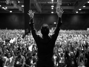 A person raises hands on stage facing a large cheering crowd in an indoor venue. The crowd's hands are also raised, creating an energetic atmosphere.