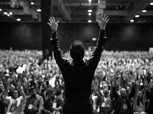 A person raises hands on stage facing a large cheering crowd in an indoor venue. The crowd's hands are also raised, creating an energetic atmosphere.