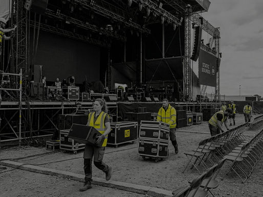 An outdoor concert stage is being set up in a large open space. The image is in greyscale, with bright yellow highlights on the crew's high-visibility safety vests. In the foreground, a woman carries a speaker while a man pushes a flight case. To the right, other crew members are carefully lining up rows of folding chairs on the gravel ground. In the background, a massive stage structure is under construction with lighting rigs, scaffolding, and large black screens, one of which displays the text "STATIC SOUNDS." The sky is overcast, and the overall atmosphere is one of focused activity and preparation.