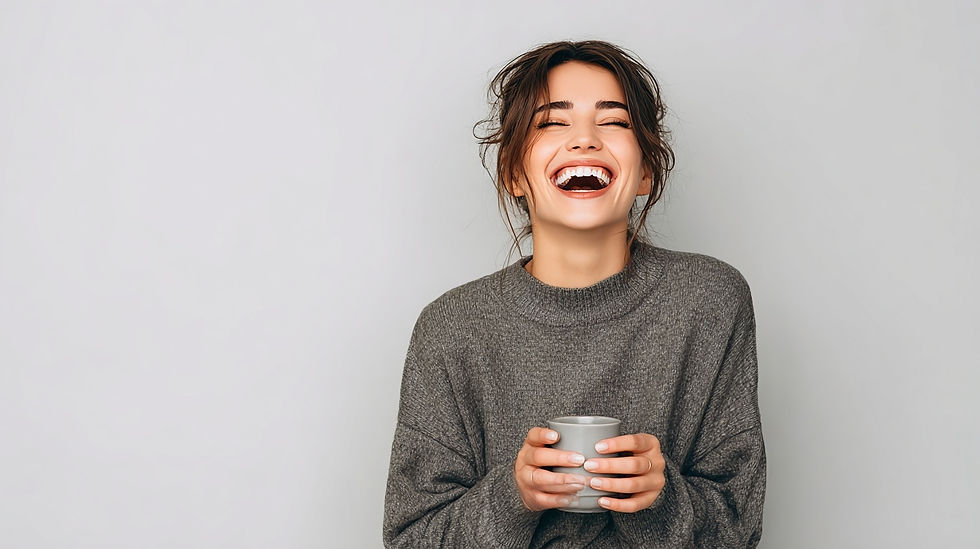 Woman in a gray sweater laughing joyfully, holding a mug. Neutral background, her hair is up, and the mood is cheerful and relaxed.