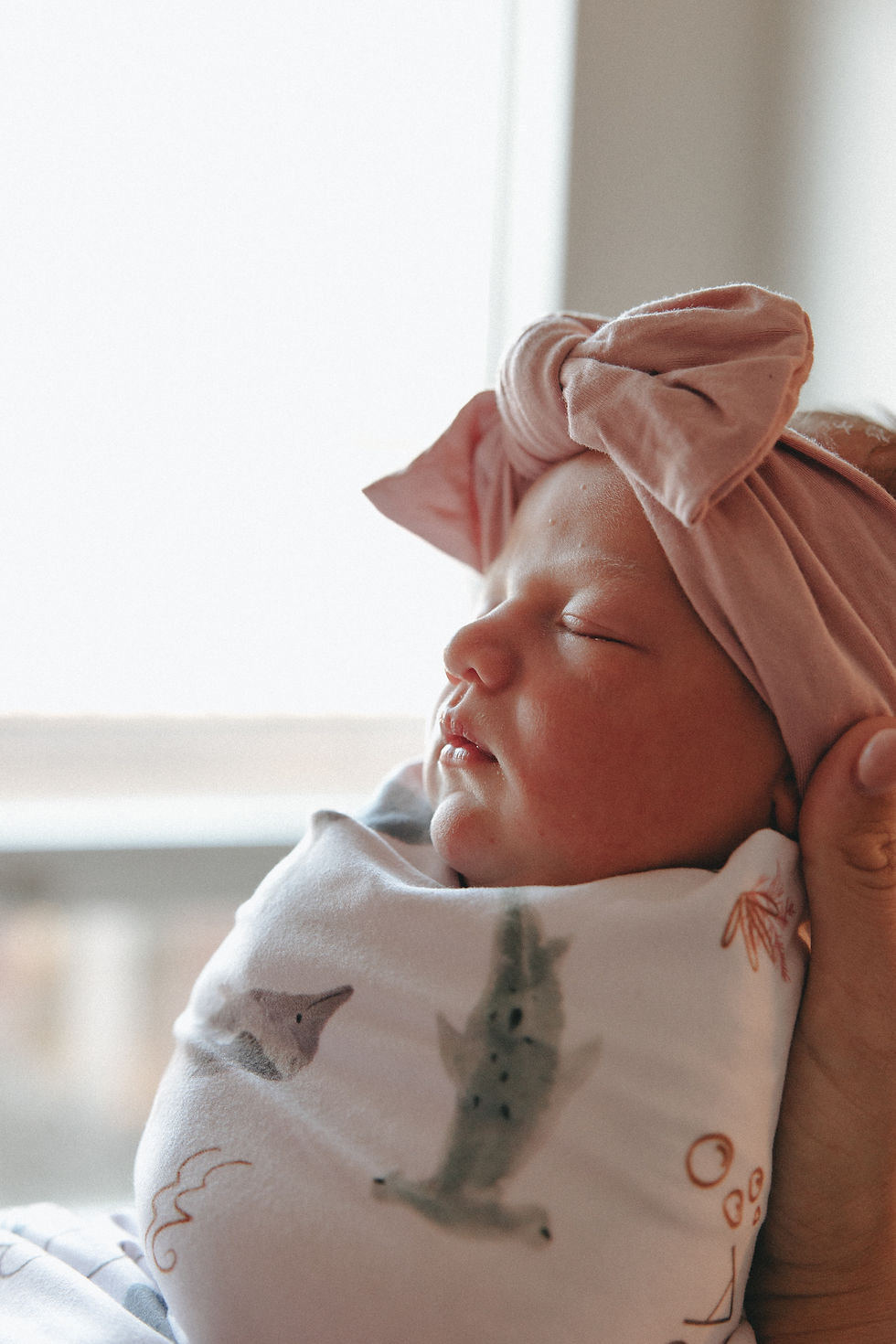Newborn swaddled in a white blanket with animal prints, wearing a pink bow headband, peacefully sleeping in a softly lit room.