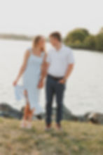 A smiling couple holds hands by a serene lake. She wears a light blue dress; he wears a white shirt. Lush greenery in the background.