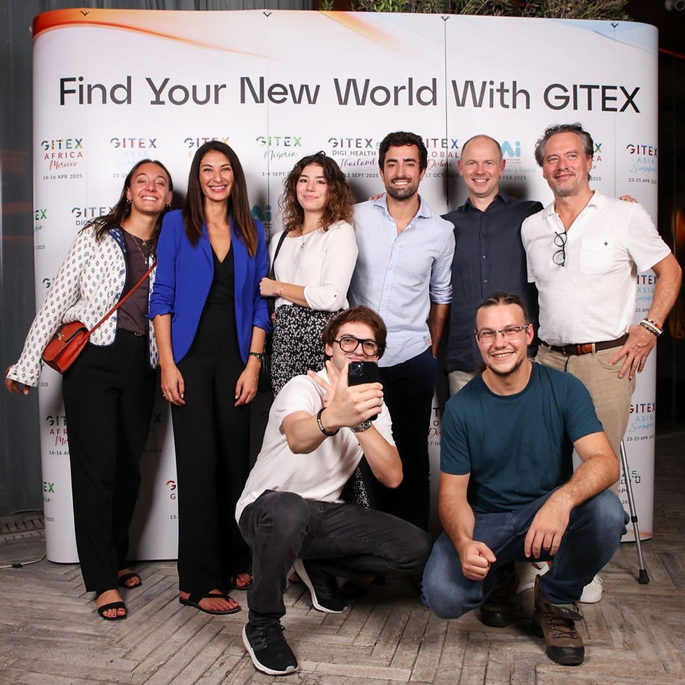 Group of eight smiling people pose for a photo. Background has text "Find Your New World With GITEX." Casual setting.