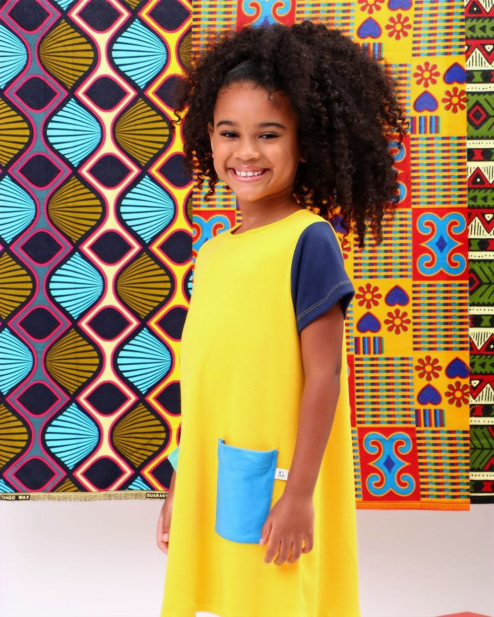 Menina negra com vestido amarelo e bolsos azuis.