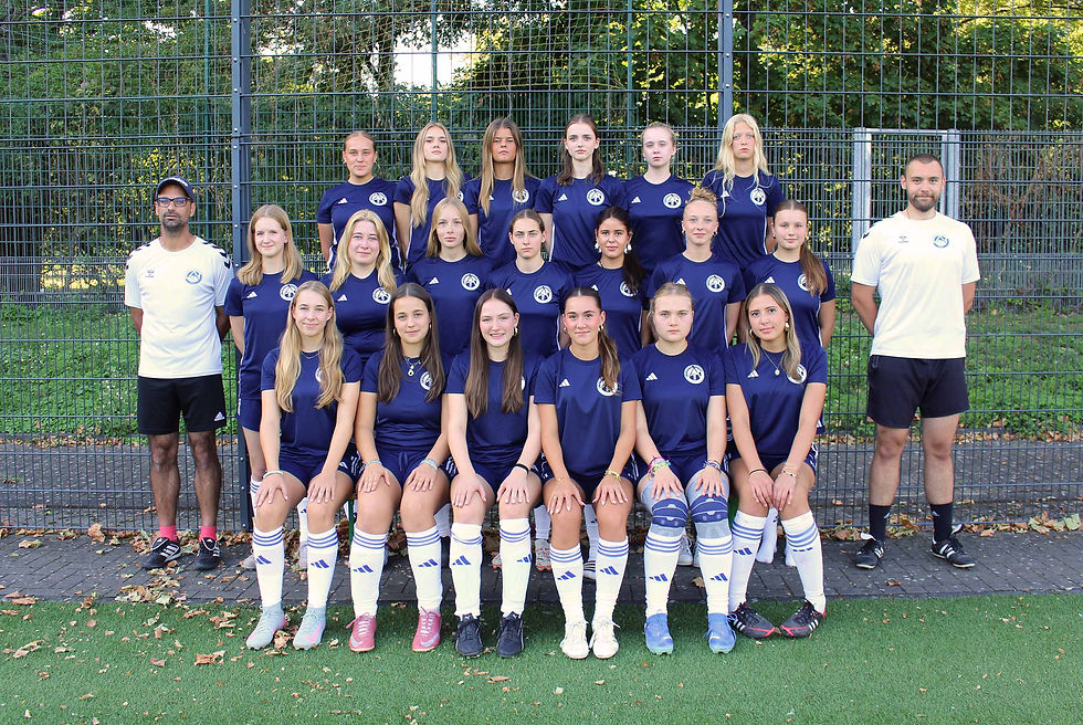 Fußball-Mädchenteam mit zwei Trainern in blauen Trikots vor Zaun, posiert ernst für ein Mannschaftsfoto.