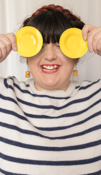 Photo of me a young plus size woman with brown hair in a braid ontop of my head with burger earrings and yellow toy plates over my eyes