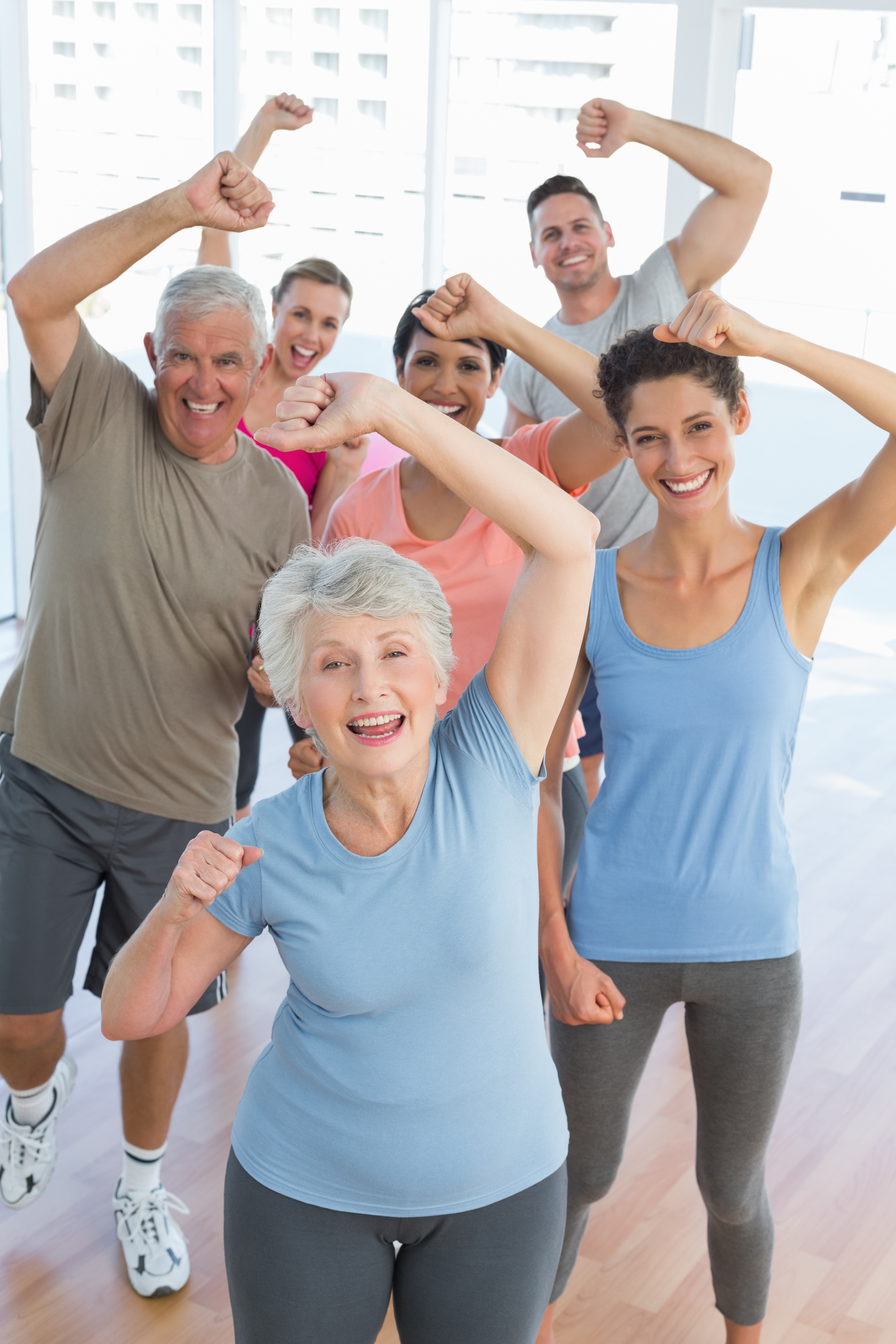 Smiling group of diverse adults in bright workout clothes doing exercises with raised arms in a sunlit room with wood floors.