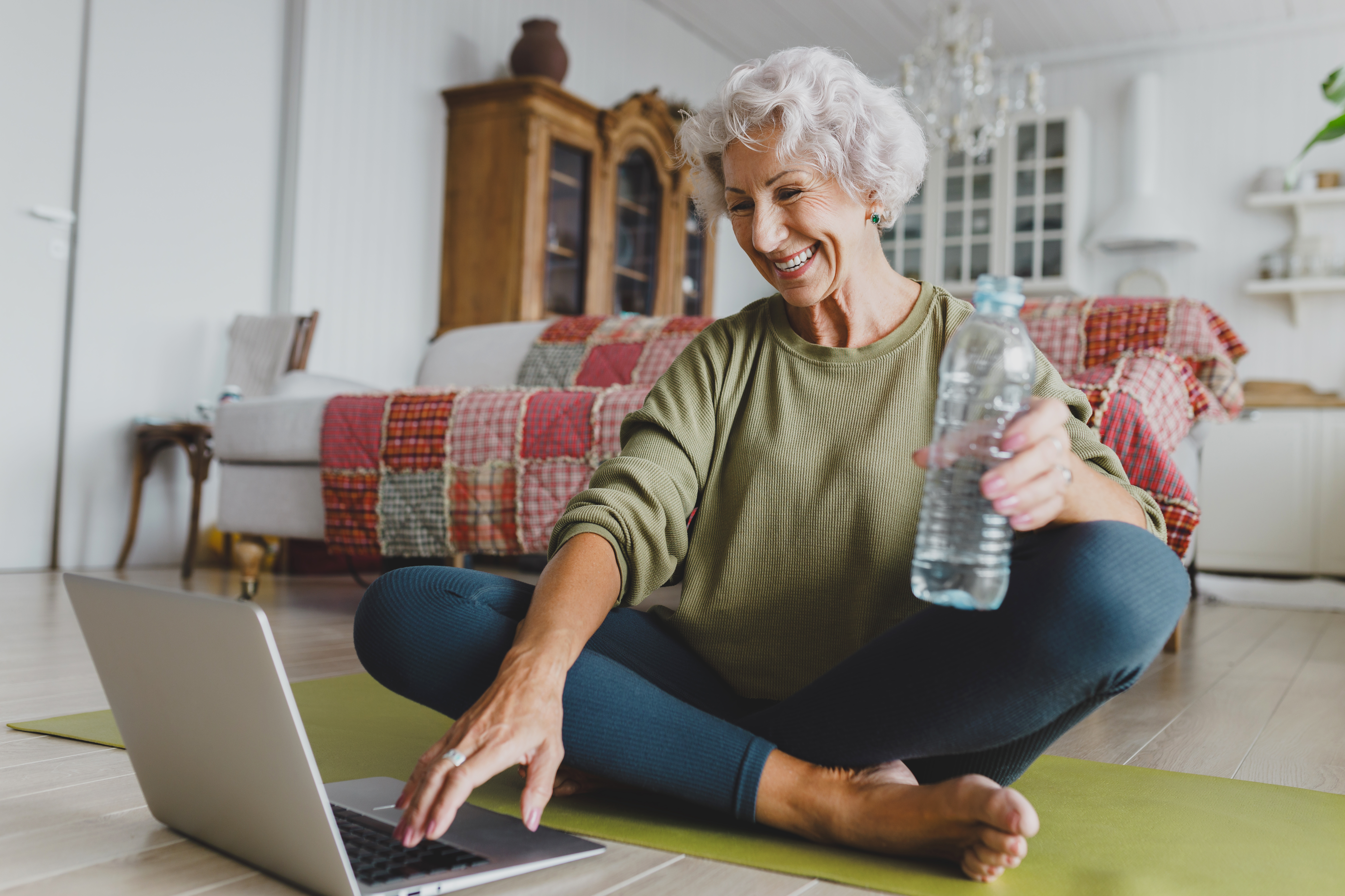 Elderly woman smiling, sitting on a yoga mat in a cozy living room, using a laptop, and holding a water bottle. Bright, inviting atmosphere.