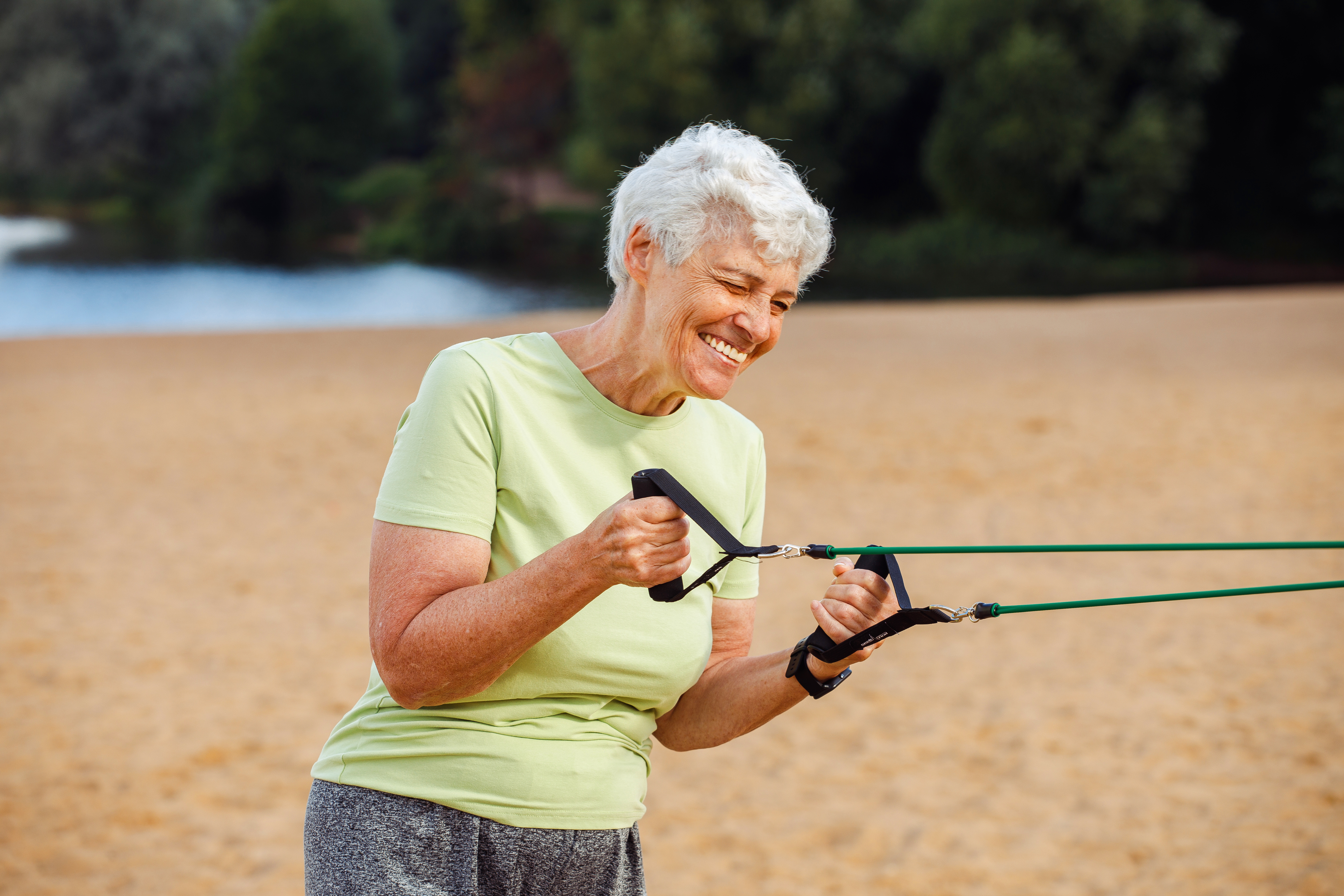 Smiling person in a light green shirt exercises with resistance bands on a sandy beach with trees in the background.