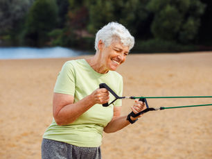 Smiling person in a light green shirt exercises with resistance bands on a sandy beach with trees in the background.