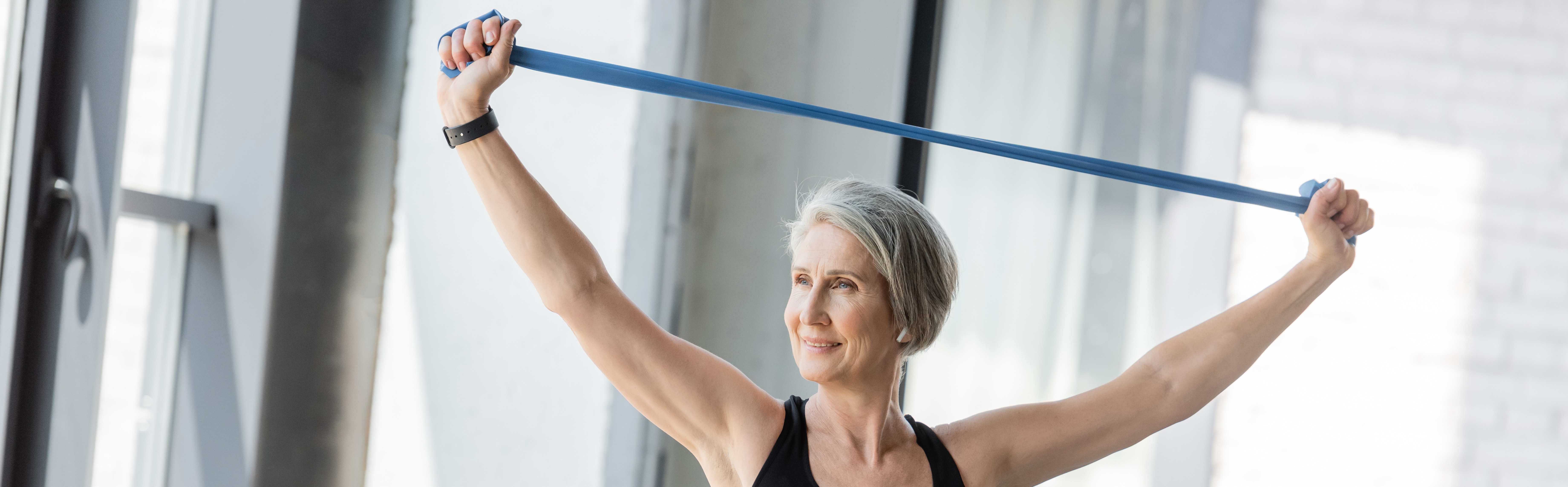 Older woman smiling while stretching a blue resistance band overhead in a bright room. Light gray walls and large windows in the background.